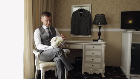 Handsome-groom-man-with-wedding-bouquet-in-his-hands-at-home-near-window-preparing-to-go-to-bride