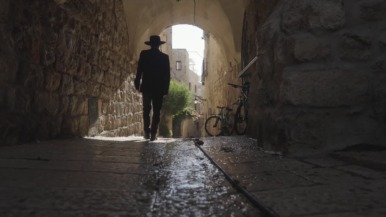 Orthodox Jewish man walking through an ancient passageway in the Old City of Jerusalem, Israel. He has side curls, black hat and suit and holds a prayer book. He is seen from behind in slow motion.