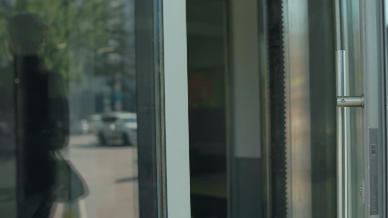 Professional banker opens glass door walking out of building with her reflection visible on door, parked cars in background, urban setting, focus on business attire, and modern environment