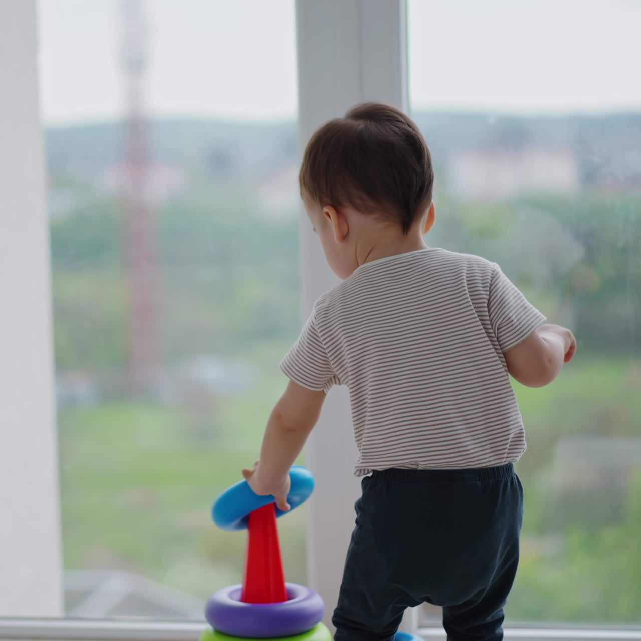 幼児の男の子がパノラマの窓の近くにあるカラフルなピラミッドに近づいてきます。元気で活発な子供がおもちゃを組み立てています。