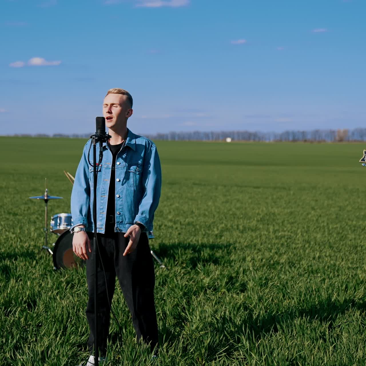 Music group performance in open air. Blond guy soloist singing into microphone while musicians playing the acoustic guitar and drums on field background
