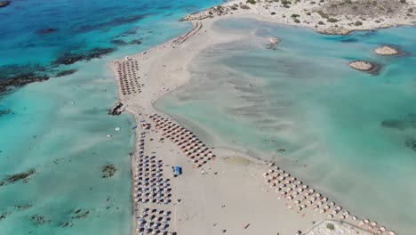 drone view in greece flying over elafonisi white sand narrow beach, clear blue water on the sides and many umbrellas on a sunny day