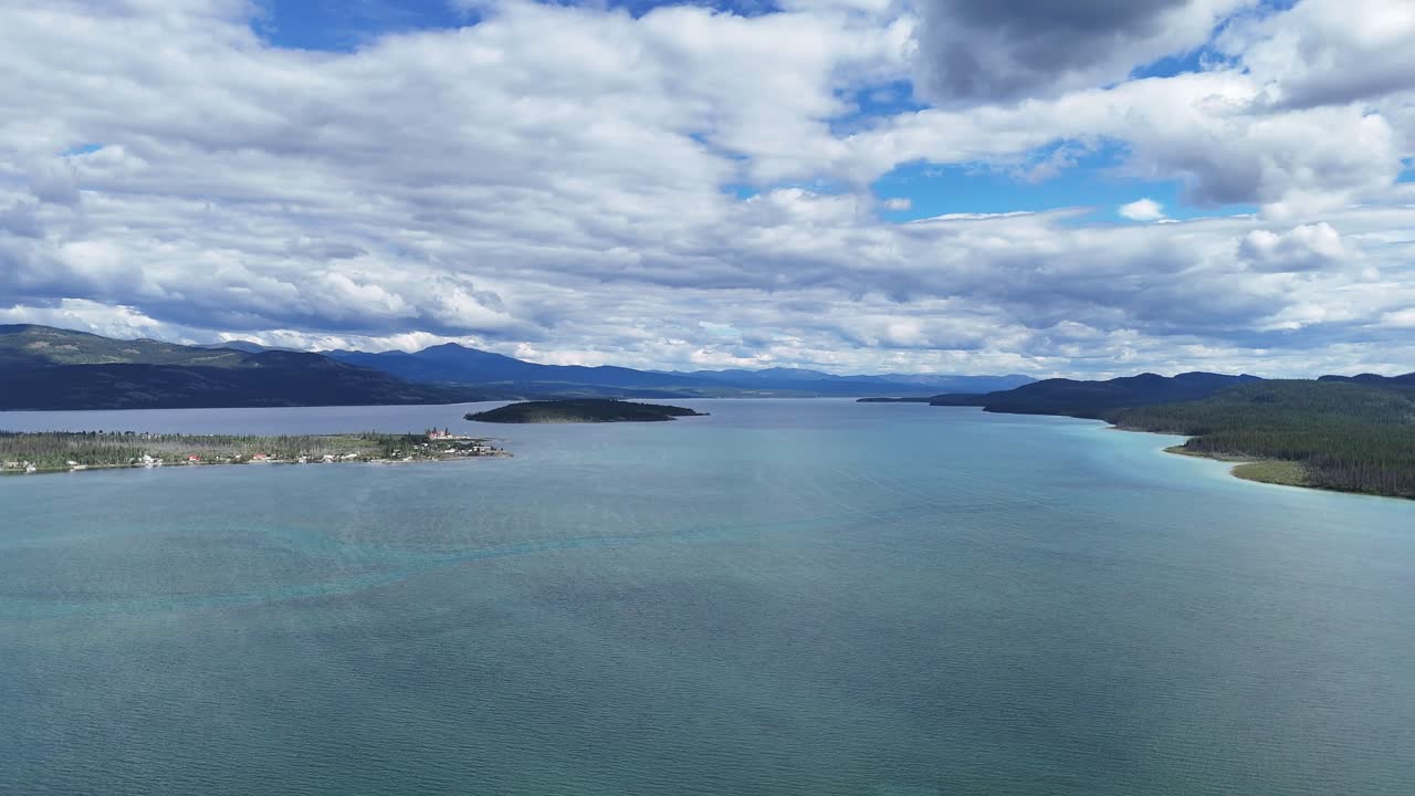An aerial view captures a vast, tranquil lake stretching towards distant, hazy mountains under a dynamic sky filled with fluffy white clouds, highlighting a serene natural landscape in canada