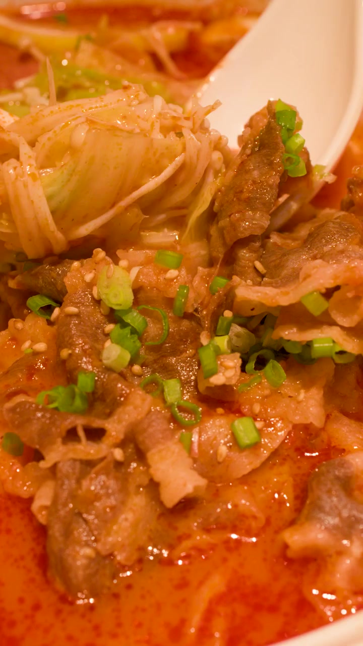 Bidikan close-up sumpit dan sendok mengangkat daging sapi dan mi dari kuah merah pedas.