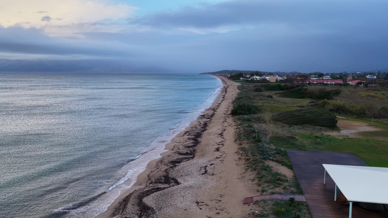 Long Sandy Coastline Aerial: Moody Sunrise or Sunset Drone View Over Tranquil Ocean Waves Breaking on Wide Beach. Mediterranean Seascape Panorama with Coastal Village in Distance and Overcast Sky