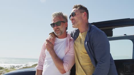 happy caucasian gay male couple wearing sunglasses embracing by car on sunny day at the beach