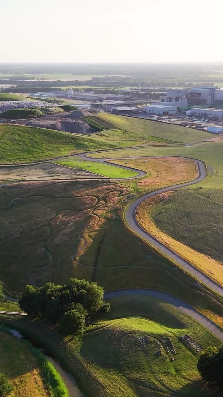 Aerial view of a winding path through green and brown hills with an industrial complex in the background