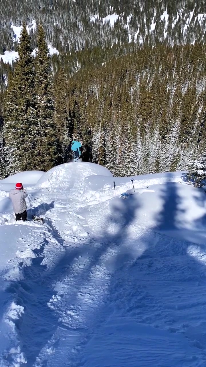 Snowboard professionistico nel backcountry: salto da scogliera su cuscino di neve fresca con Indy grab in aria. Inverno a Vail Pass, Colorado. Reel verticale delle Montagne Rocciose con neve profonda. Snowboarder che filmano trick con lo snowboard sotto un cielo azzurro in una mattina soleggiata.