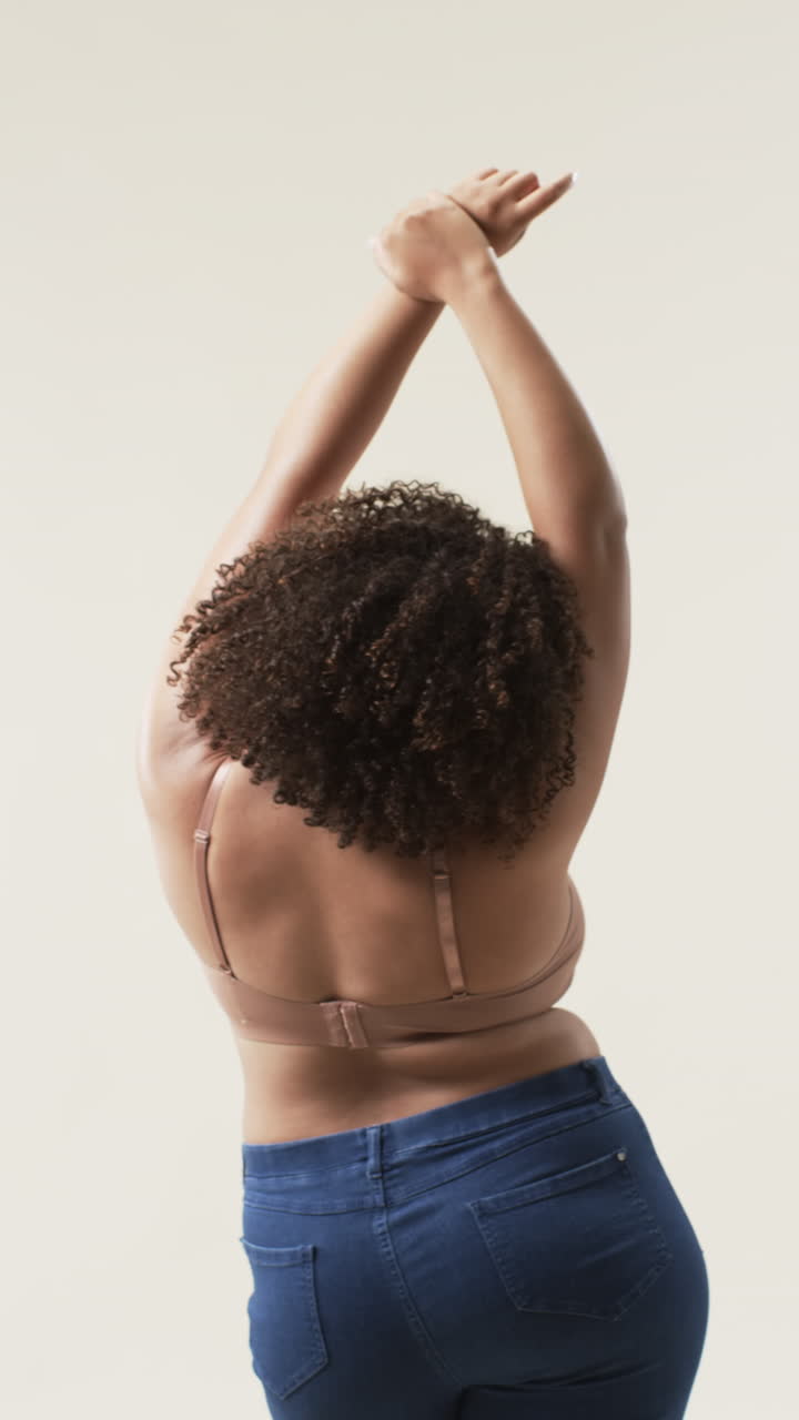 Vertical video: Stretching arms, plus size woman in jeans and bra promoting body positivity