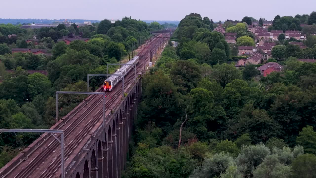 Drone reverses along railway track at Digswell Viaduct as a train moves forward at dusk. The viaduct is framed by green trees, houses and soft evening light