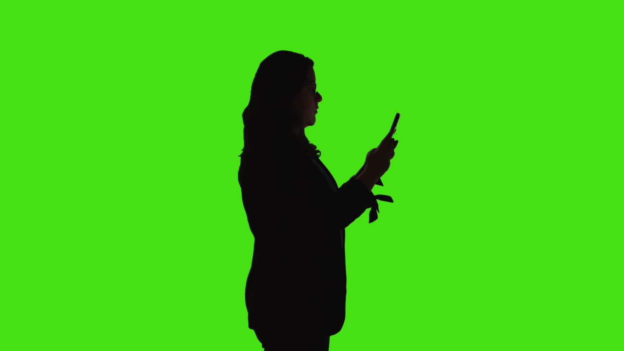 낮은 키 조명으로 녹색 화면에 서서 문자 메시지를 보내거나 읽거나 휴대 전화로 온라인을 스크롤하는 사업가의 실루