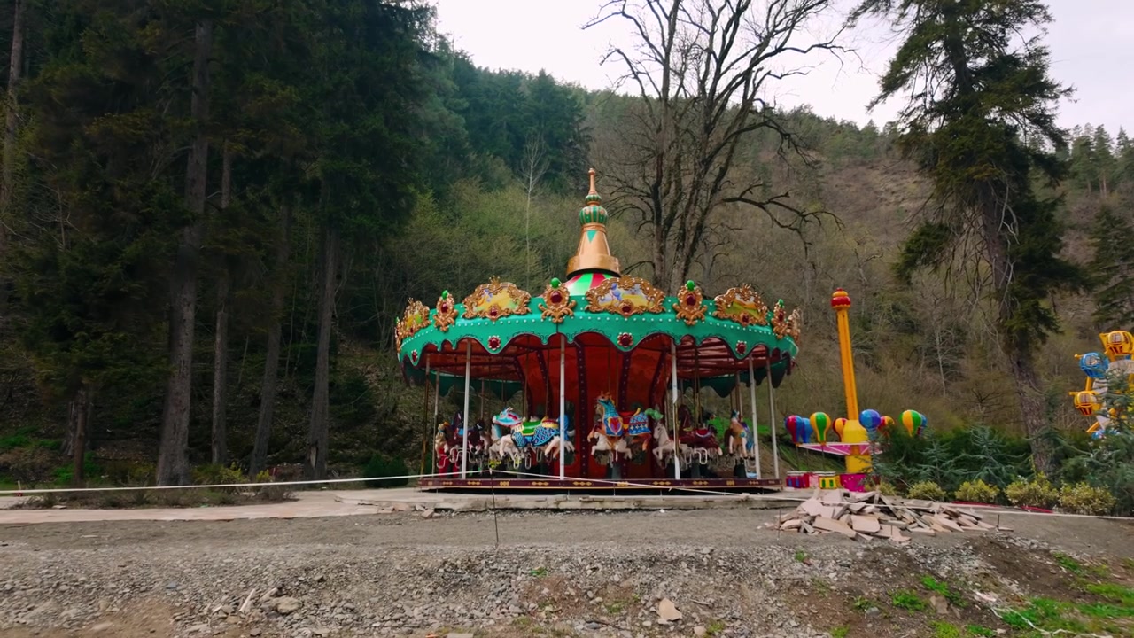Yumuşak bulutlu bir gökyüzünün altında, yemyeşil ağaçlar ve ormanlık tepelerle çevrili bir park ortamında renkli vintage atlıkarınca ve diğer küçük eğlence parkı oyuncakları.