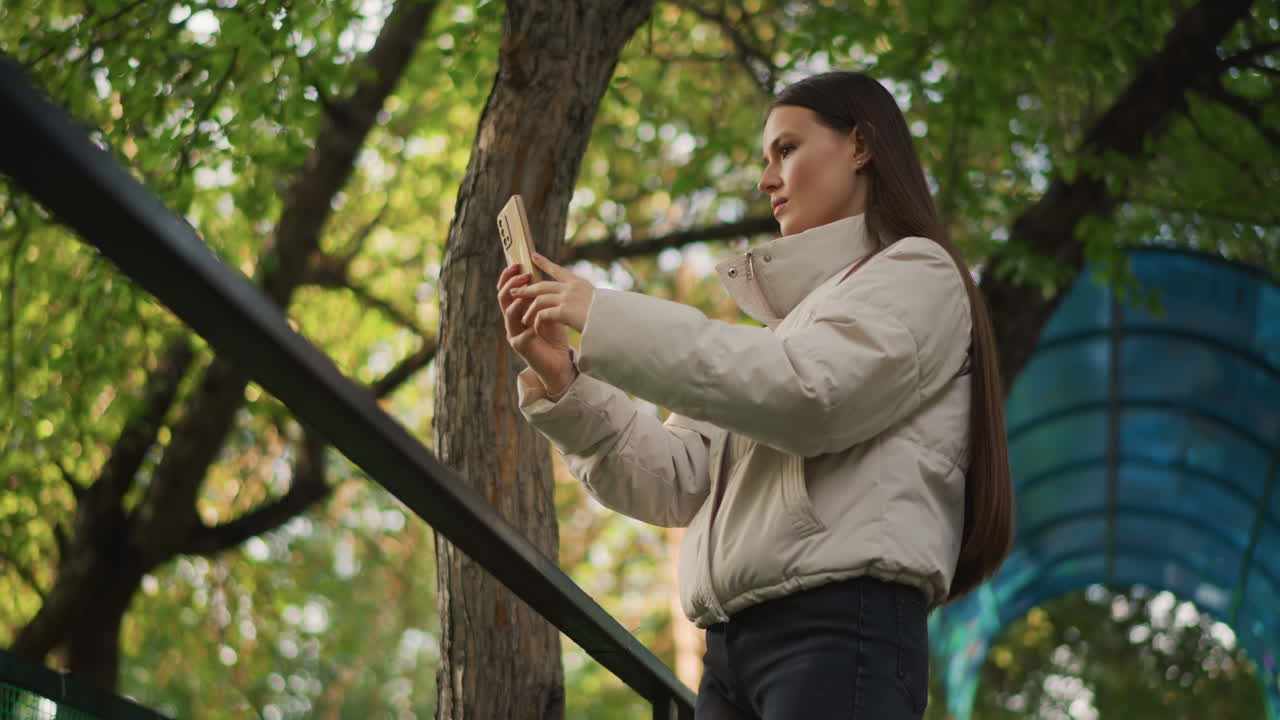 Donna caucasica che si scatta un selfie all'aperto in un parco, indossa un piumino beige e jeans, in piedi accanto a una ringhiera nera sotto una tettoia verde e un camminamento blu, espressione concentrata, mentre usa uno smartphone