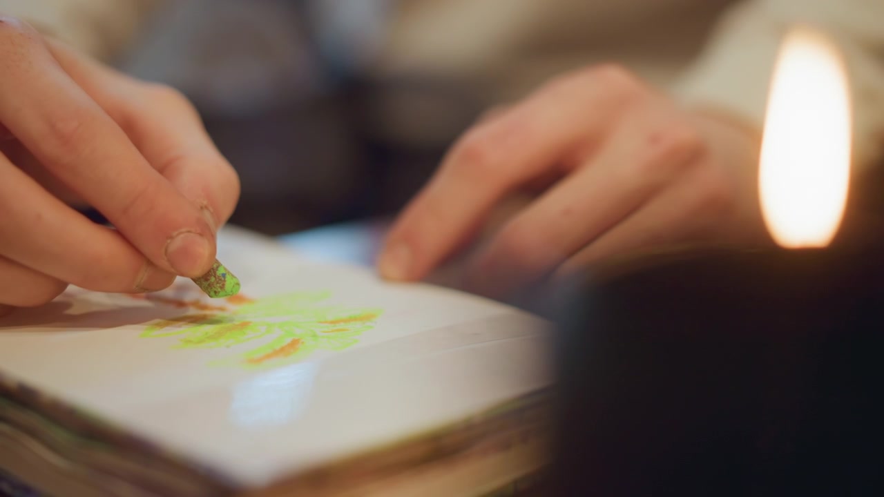 Close up of hands drawing with green and orange crayon on white paper in sketchbook. Hands move steadily creating colorful abstract lines under soft lighting from candle flame. Background