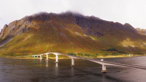 Aerial-view-of-a-white-camper-van-crossing-a-gorgeous-bridge-in-the-fjords-of-the-Lofoten-Islands,-Norway