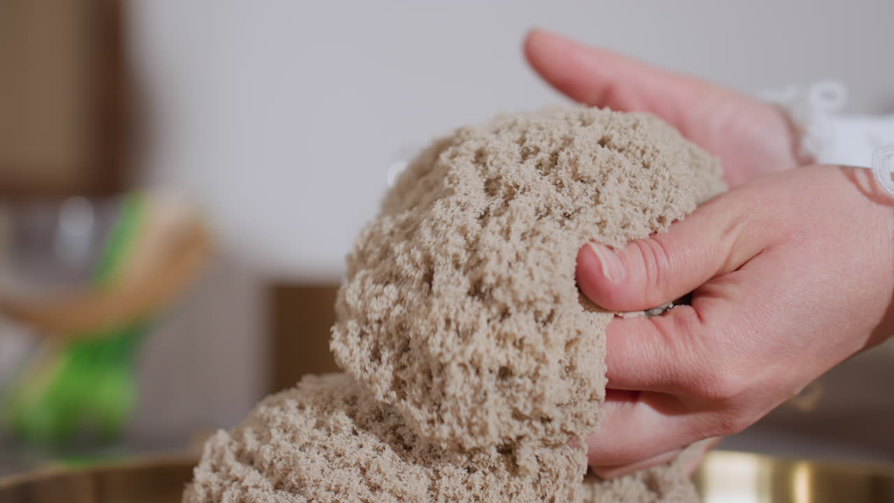 Side view of therapy patient gently lifting and smoothing soft wet soil mold with both hands during calming sensory therapy session, showing tactile interaction on golden tray with blurred background