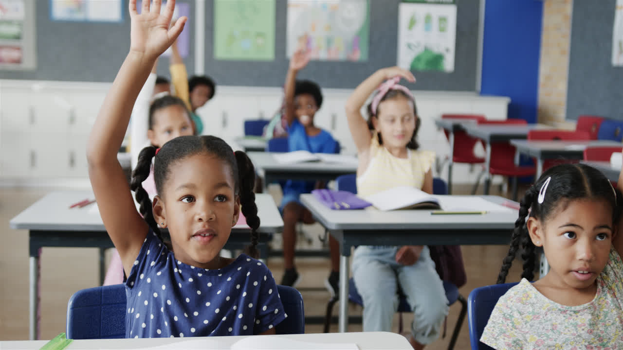 Happy diverse schoolchildren at desks raising hands in elementary school classroom in slow motion