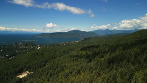 drone flying forward over the confer tree forested mountain side of west vancouver, canada on a sunny summer day with blue ocean water and bowen island in the distance