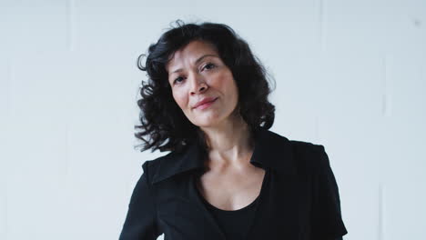 Portrait-Of-Smiling-Mature-Businesswoman-In-Suit-Walking-Into-Focus-Against-White-Studio-Wall