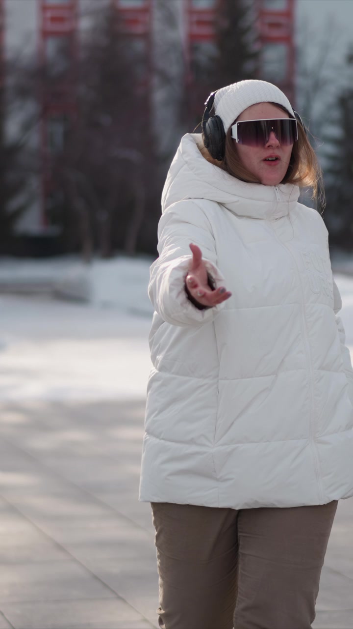 Woman wearing white puffer jacket black headphones and sunglasses dances outdoors in winter. Woman moves her arms expressively while walking on snow covered ground with bare trees