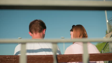 mujer con gafas apoyadas en la cabeza interactúa con el hombre sentado estrechamente en un banco al aire libre, bajo un cielo brillante y enmarcado por barandillas de hierro