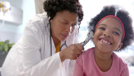 African-american-female-doctor-examining-ear-of-happy-girl-in-hospital-room,-slow-motion