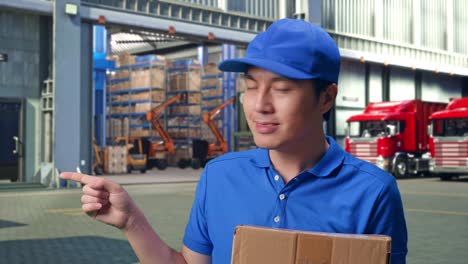 Close Up Of Asian male Courier In Blue Uniform Smiling And Pointing To The Side While Delivering A Carton, Outside of Logistics Distributions Warehouse