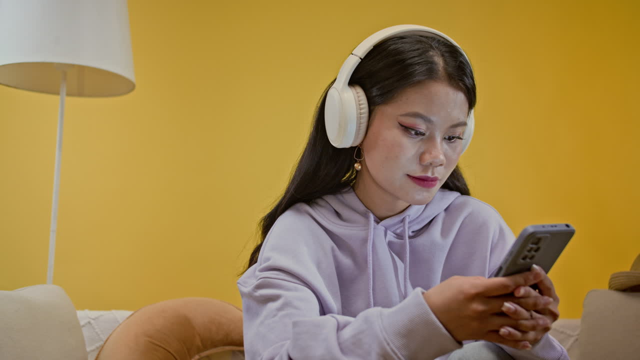 Asian Teenage Girl in Headphones Texting on Smartphone on Yellow Background