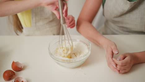 partial view of girl in yellow apron whisking flour and egg mixture in glass bowl while woman sits beside with hands folded, broken eggshells scattered and flour stains spread across table surface