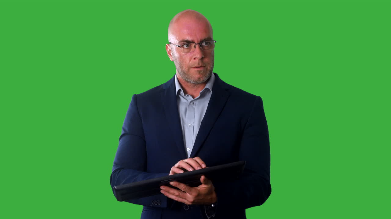 A bald business man in a suit with a green screen background, holding and typing on a computer keyboard