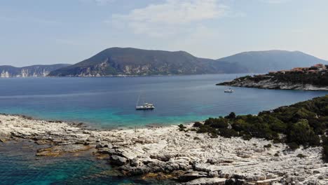 Ruhiges-Türkisfarbenes-Und-Blaues-Wasser-An-Einem-Steinigen-Abgelegenen-Strand-Mit-Booten,-Die-Im-Meer-Von-Paralia-Emplisi-Erisos-Griechenland-Verankert-Sind---Luftaufnahme-Mit-Vorwärtsschwenk