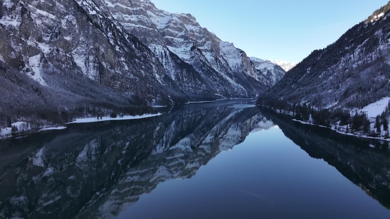 Drone pushes towards Klöntalersee perfect reflections of snow covered alps, serene winter escape in Switzerland's natural beauty.