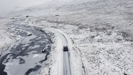 Faroe-Islands,-4K-Aerial-tracking-behind-black-car-in-the-snow-with-beautiful-mountains-in-the-background