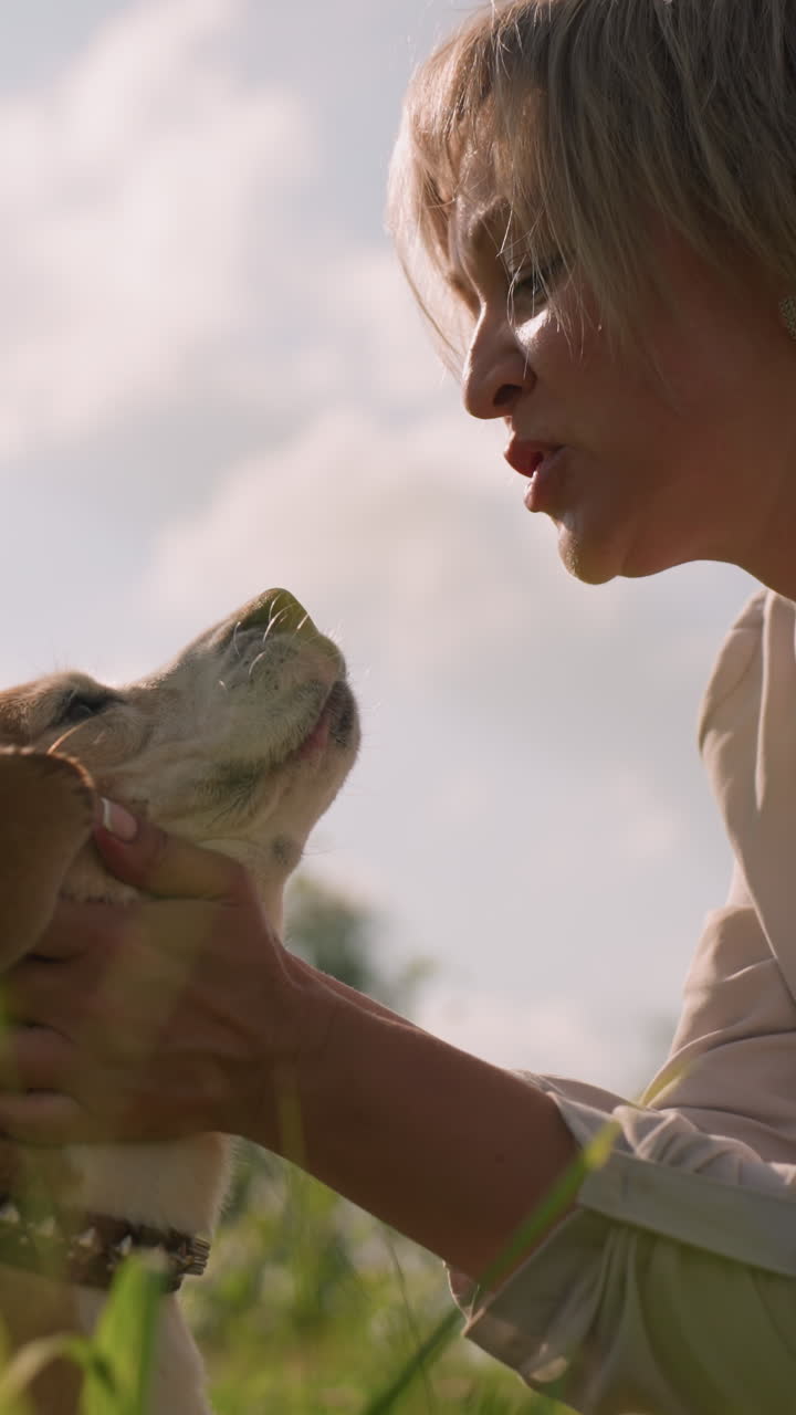 Woman holding dog's head lovingly brings face close as dog playfully licks her mouth and his nose in grassy field under bright sky, affectionate interaction showcasing strong bond
