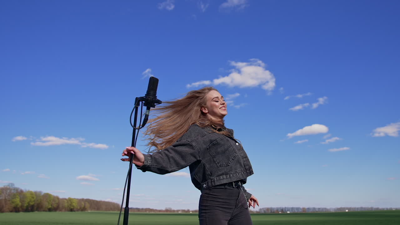 Smiling girl singing into a mic among nature. Attractive young female singer performing a song and dancing happily on green field under blue sky. Slow motion.