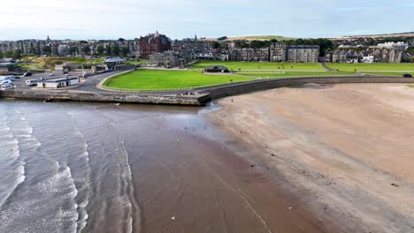 Drone footage moves steadily over sandy shoreline toward Victorian architecture, green golf course, and coastal town under bright daylight with soft shadows