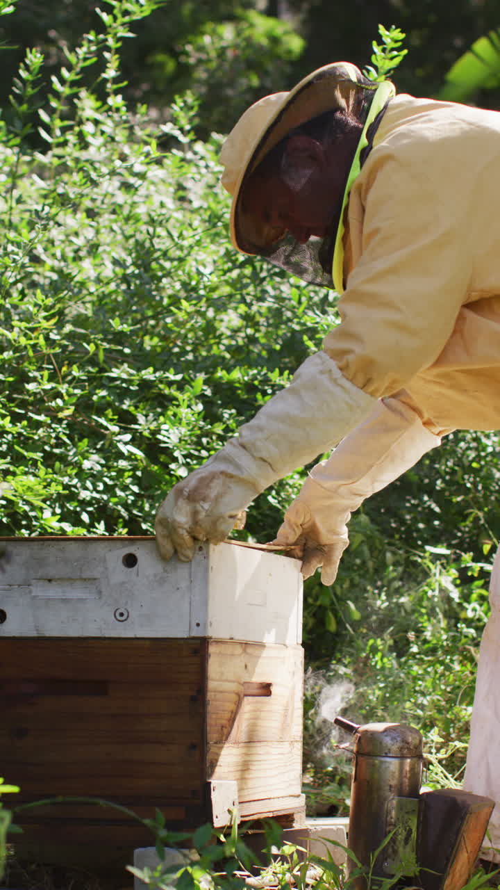 fotografía vertical de un apicultor caucásico de edad avanzada abriendo una colmena, con ropa de protección