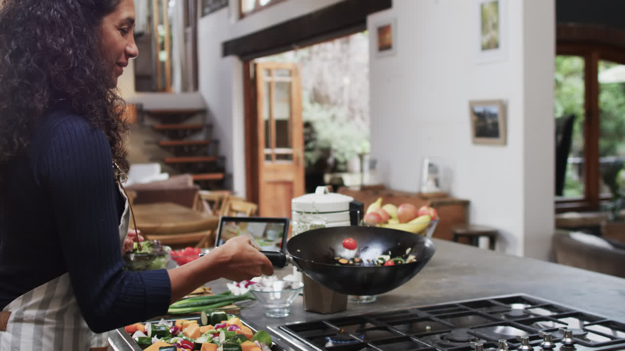 mujer biracial feliz preparando comida usando tableta en la cocina, cámara lenta con espacio de copia