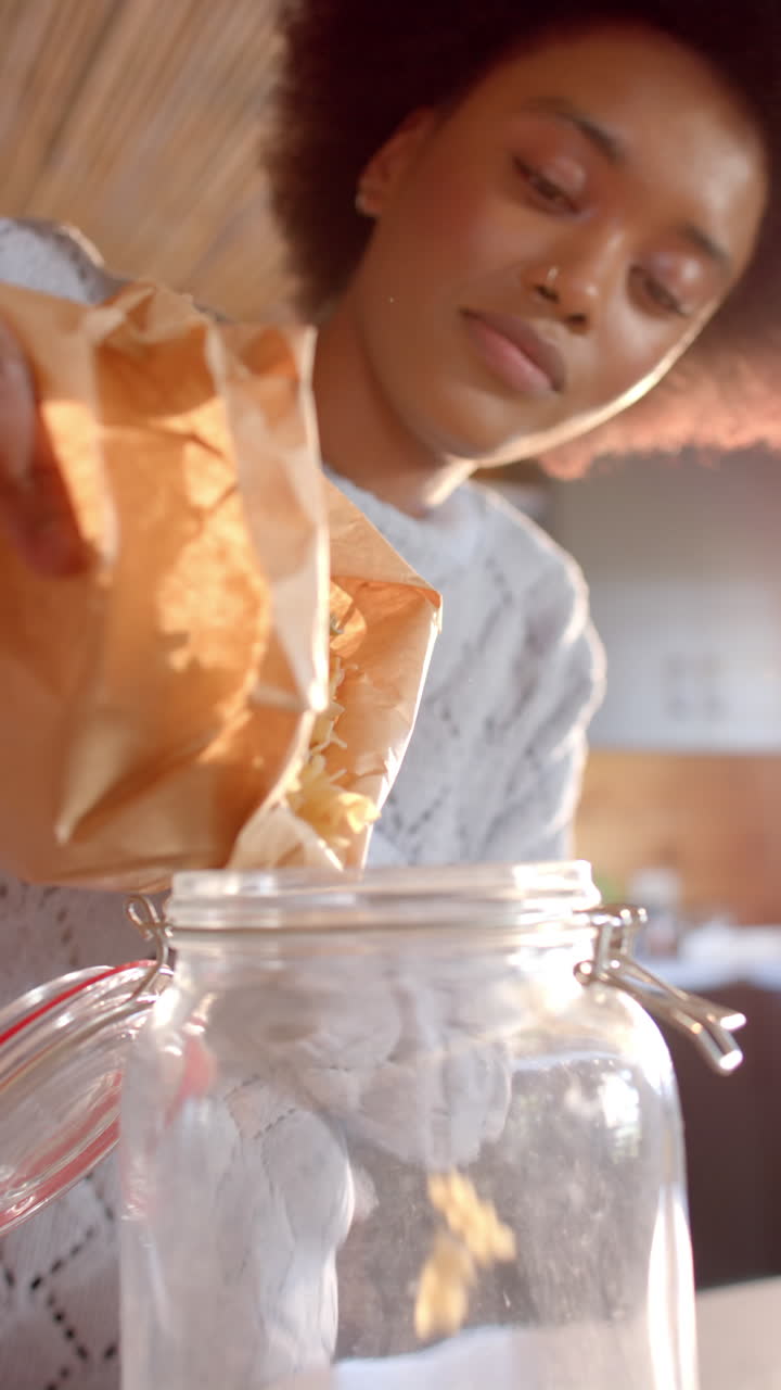 Vertical video of african american woman pouring pasta into jar in sunny kitchen, in slow motion
