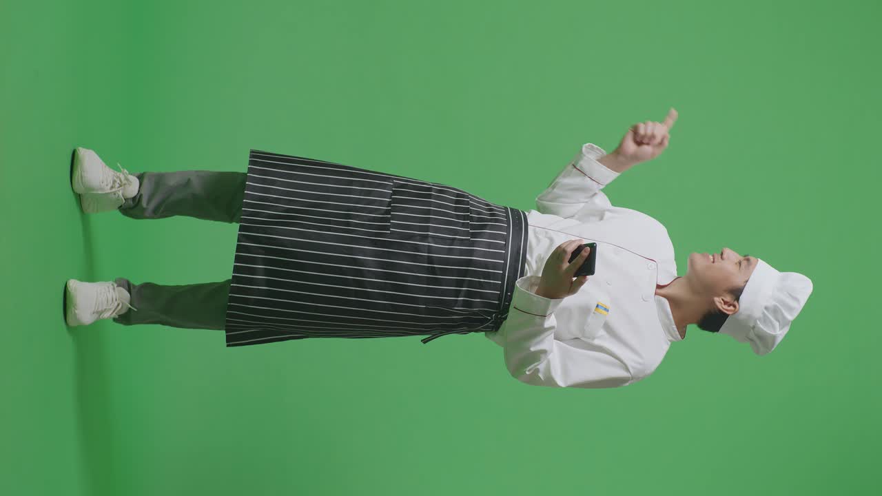 녹색 스크린 배경 스튜디오에 서 있는 동안 스마트폰을 사용하고 을 가리키는 아시아 남자 셰프의 전체 몸