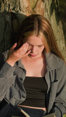 la dama se sienta bajo el árbol leyendo un libro mientras se apoya en el tronco, se frota la ceja y se ajusta el cabello, apareciendo pensativa, la luz del sol ilumina suavemente su rostro, con las hojas balanceadas por el viento