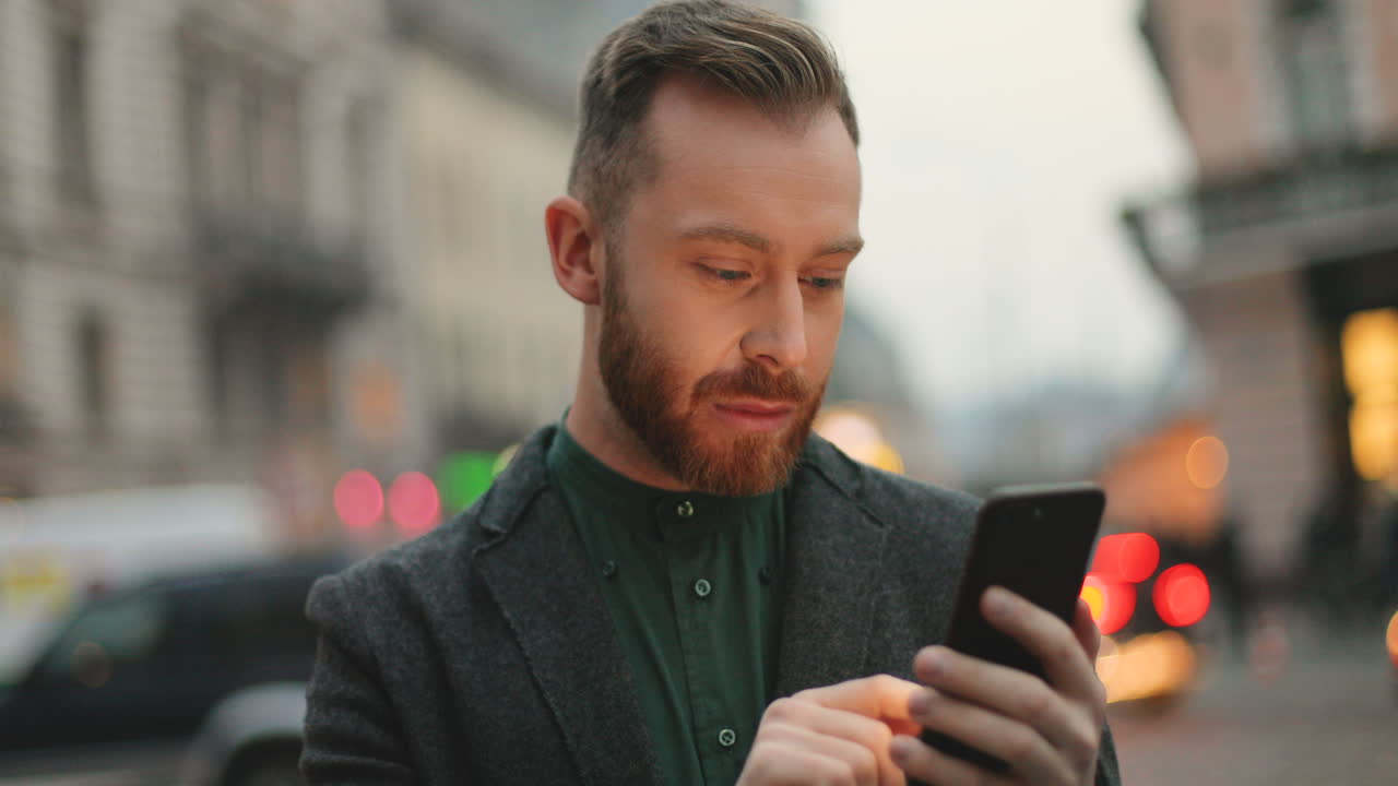 Close-up view of caucasian businessman with a beard texting on the phone in the street with city lights of the background