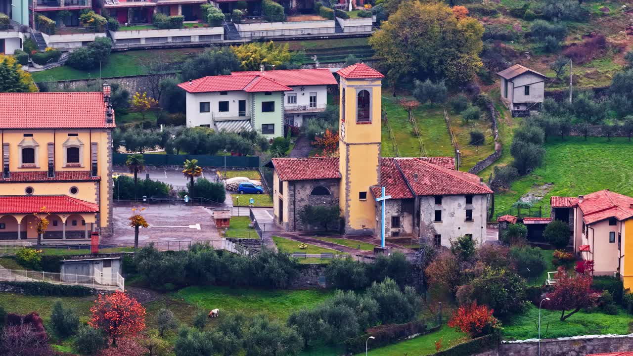 Vista aerea di un piccolo villaggio vicino al Lago d’Iseo, che mette in mostra un campanile rustico giallo, case tradizionali, uliveti e verdi colline in un pacifico contesto rurale italiano