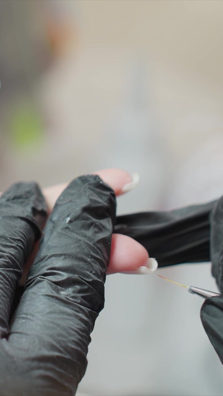Gloved fingers receive detailed nail art as another gloved pair uses fine tools for precise design. focus remains on meticulous hand movements and specialized equipment highlighting skill