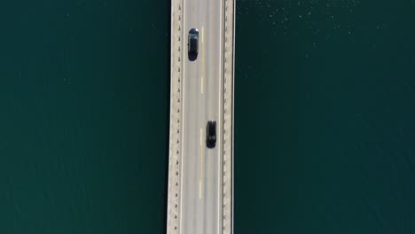 Birdseye-aerial-view-of-left-hand-road-traffic-on-bridge-above-river-water,-top-down-drone-shot