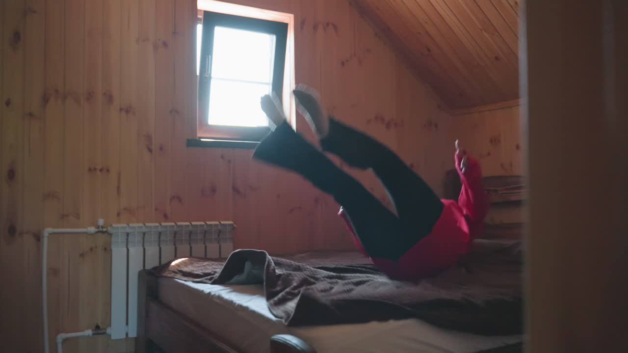 young girl in red outfit preparing to jump on bed in wooden cabin room as soft daylight streams through window, cozy atmosphere and back view create relaxed, playful mood in peaceful indoor setting