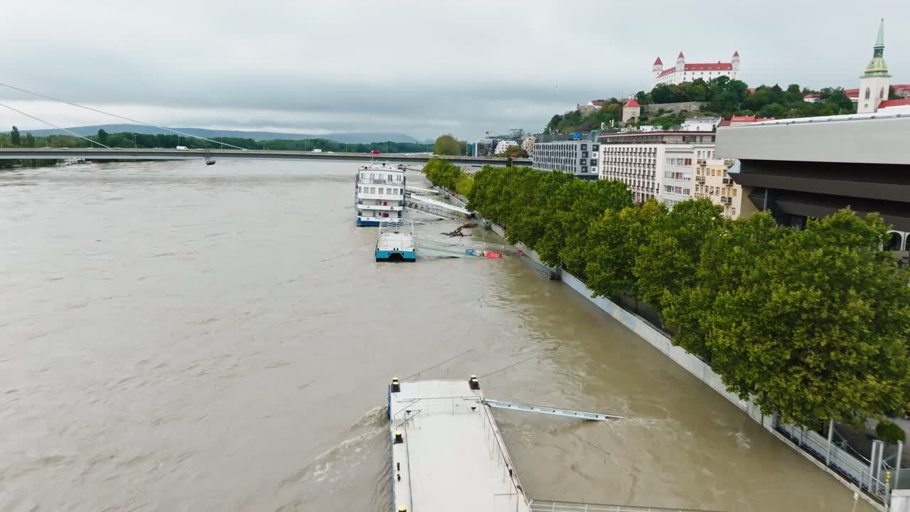 Aerial view of flooding in Bratislava, showcasing the overflowing Danube River, submerged trees, and affected infrastructure. The city landscape and iconic landmarks are visible. (4K)