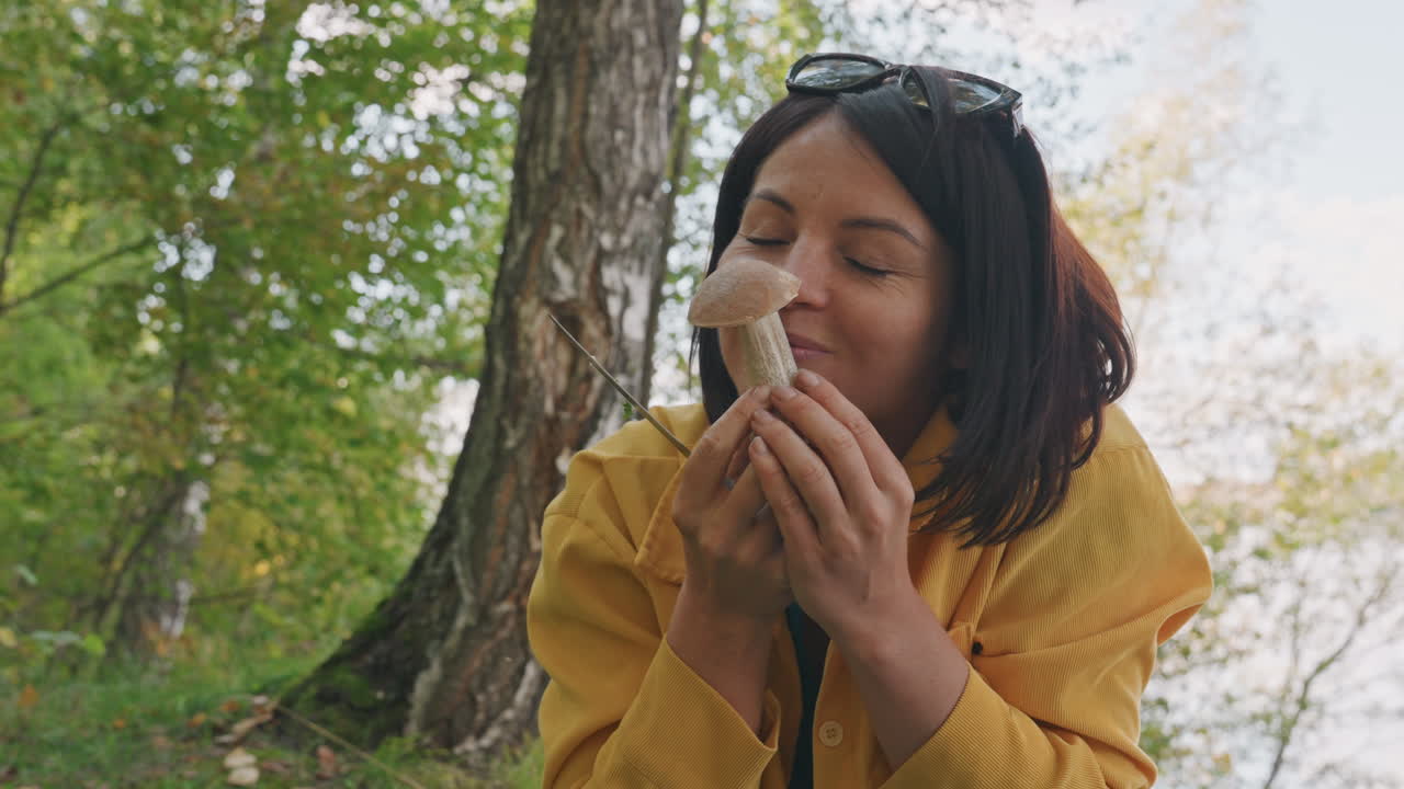 Una donna in campeggio esamina da vicino un fungo selvatico nel bosco, annusandone il cappello e sorridendo mentre mostra l'esemplare alla telecamera sotto la calda luce del giorno. Con un tronco d'albero come sfondo, traspare una giocosa curiosità culinaria.