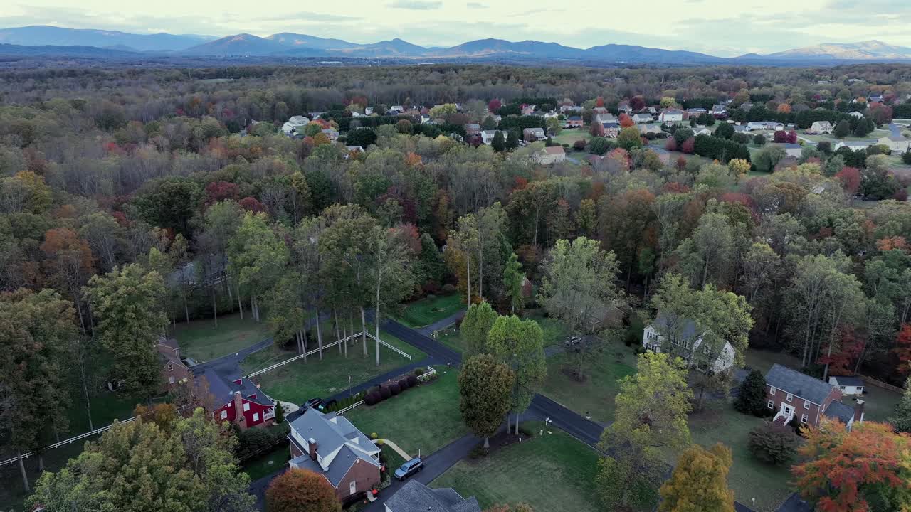 Charming street between colored trees with two-story homes in amErica suburb. Aerial wide shot. mounTain range and clouds in distance. Aerial wide shot. Sunset time in fall season. VA,USA