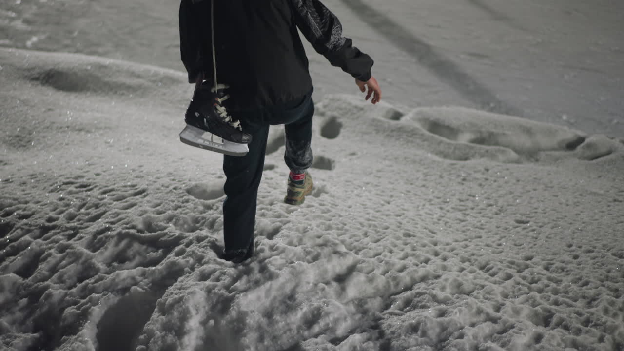 Back view of skater stepping carefully down snowy stairs holding black ice skate boot, surrounded by thick layers of snow and rough winter textures under soft lighting on cold night